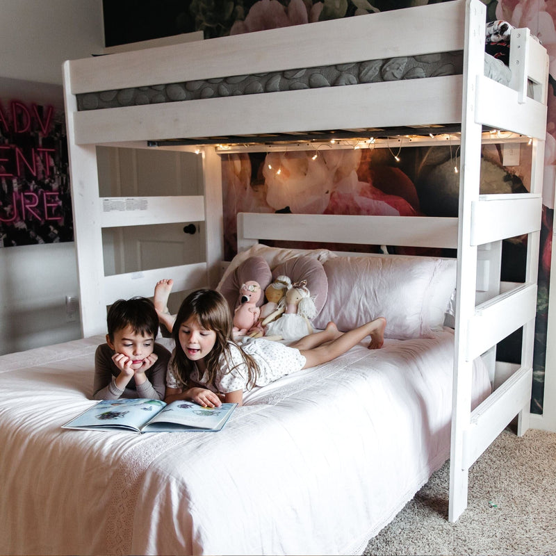Max and Lily Modern Farmhouse Twin Over Full L-Shaped Bunk Bed in White Wash - two children reading a book on the lower full bed beneath the upper bunk with string lights.