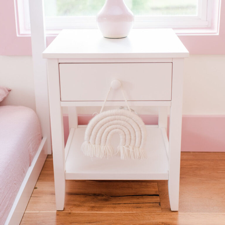 Max And Lily Night Stand With Drawer And Shelf White Room Scene 1 Max and Lily Night Stand With Drawer and Shelf in White - styled in a bedroom with a lamp on top and a woven rainbow decoration on the lower shelf beside a bed.