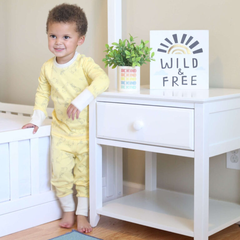 Max And Lily Night Stand With Drawer And Shelf White Room Scene 8 Max and Lily Night Stand With Drawer and Shelf in White - placed in a child's room with a plant and Wild and Free sign on top while a toddler stands nearby.