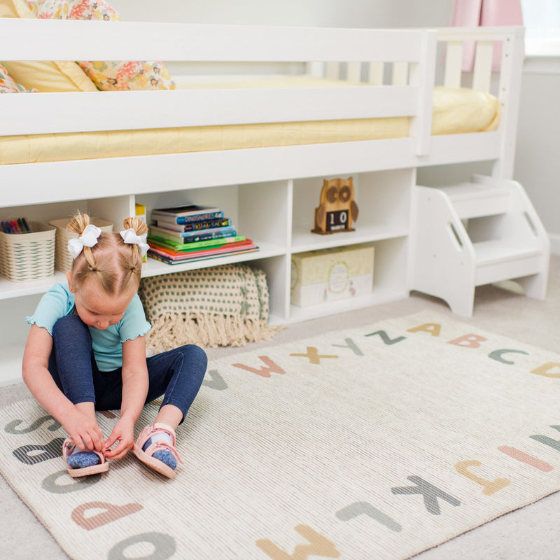 Max and Lily Soren Twin Ultra Low Loft Bed With Bookcase in white - young child sitting on an alphabet rug beside the bed's storage bookcase and step stool.