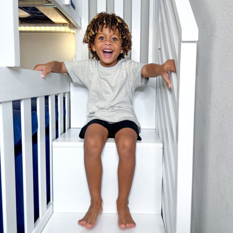 Max And Lily Twin Over Full Bunk Bed With Stairs And Storage White Staircase Detail Max and Lily Twin Over Full Bunk Bed with Stairs and Storage in White - a smiling child sitting on the wide staircase steps with under-bed LED lighting behind.
