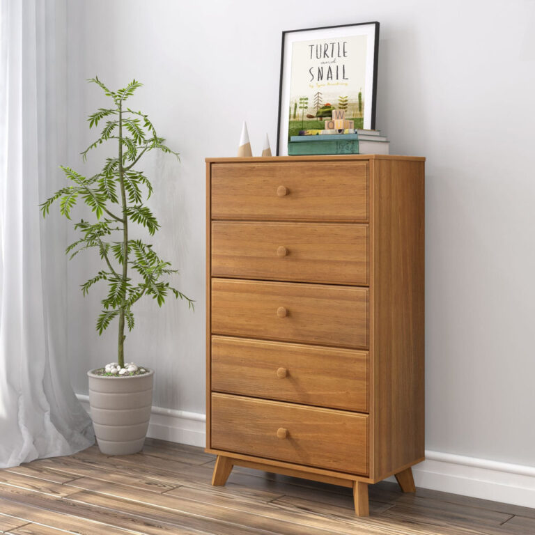 Max Lily Classic 5 Drawer Dresser Pecan Room Scene 1 Max & Lily Classic 5 Drawer Dresser in Pecan - angled view in a bedroom corner beside a potted plant and curtain with framed artwork displayed on top.