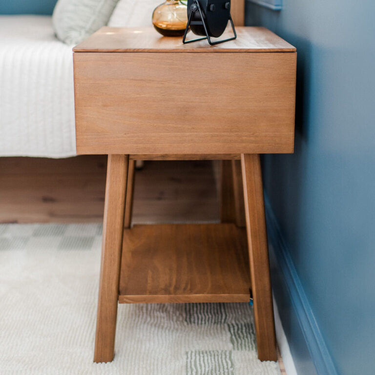 Max Lily Mid Century Modern Cubby Night Stand Pecan Side View Max & Lily Mid-Century Modern Cubby Night Stand in Pecan - side close-up beside a bed showing the wood grain and lower shelf.