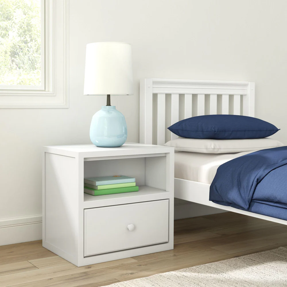 Maxtrix 1 Drawer Nightstand in White - angled bedside view in a kids room with a lamp on top, books on the open shelf and a bed with navy bedding beside it