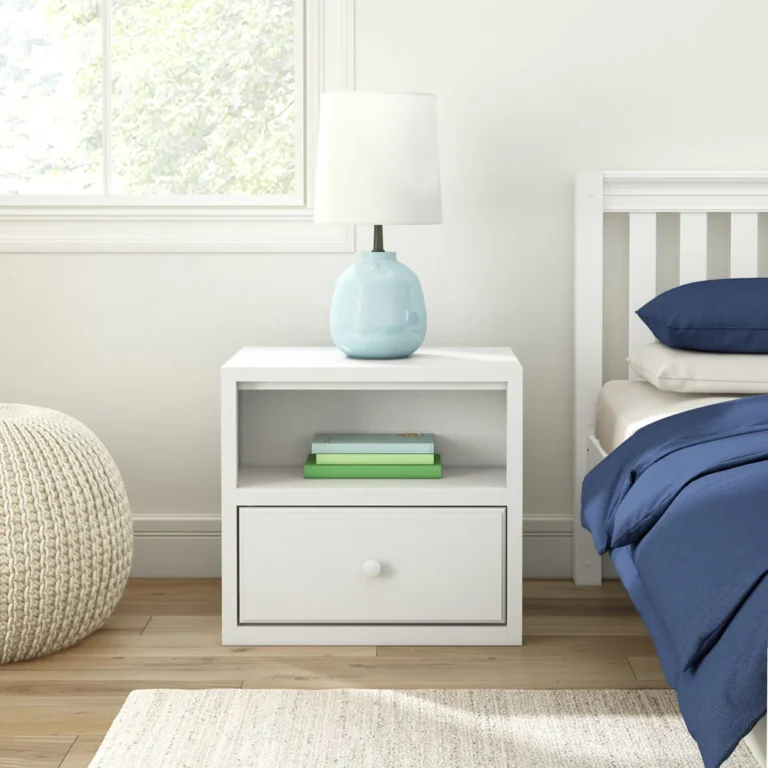 Maxtrix 1 Drawer Nightstand in White - front view beside a bed in a bright bedroom, with a lamp on top, books on the open shelf and the drawer closed