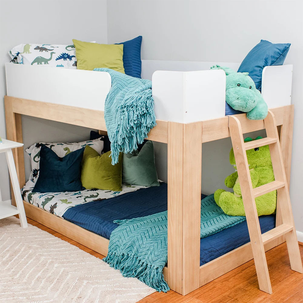 Mid-Century Modern Twin Over Twin Low Bunk Bed in White/Blonde - angled room scene with dinosaur bedding, plush toys, white safety guardrails and angled wood ladder